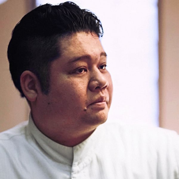 白いスタンドカラーのコックコートを着た人物の上半身クローズアップ。背景に柔らかな光が差し込む室内。