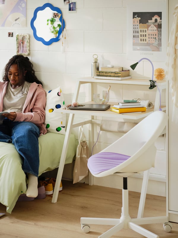 College girl sitting on bed with green bedding beside white student desk and white and purple swivel chair in white dorm room.