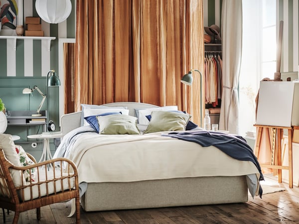 Bedroom with beige bed in the middle of the room, with orange curtains in the back of the room and green/white striped wall.