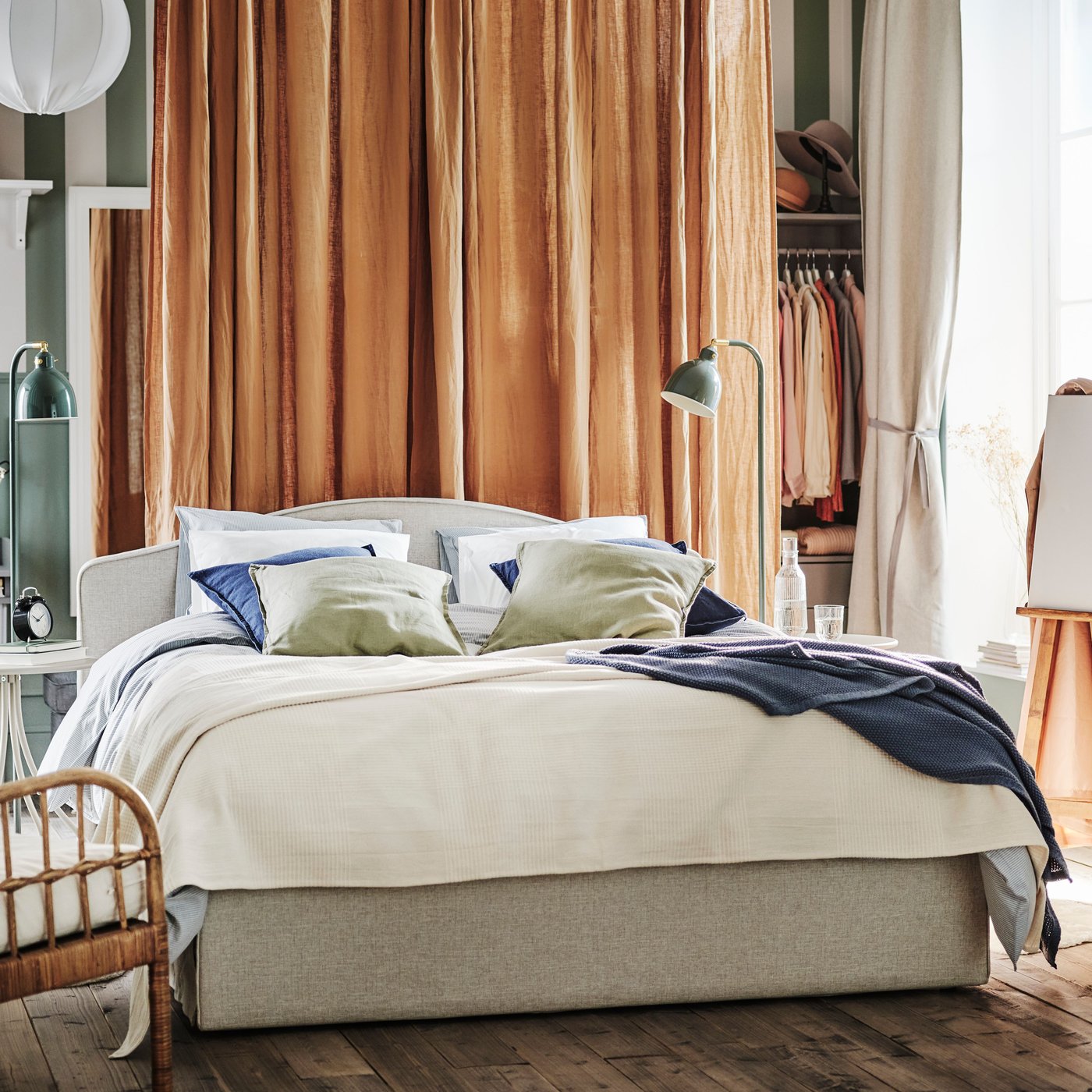 Bedroom with beige bed in the middle of the room, with orange curtains in the back of the room and green/white striped wall.