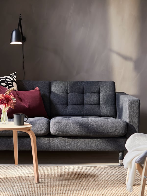 Harmaa kahden istuttava LANDSKRONA-sohva olohuoneessa harmaata auringon valaisemaa seinää vasten.
