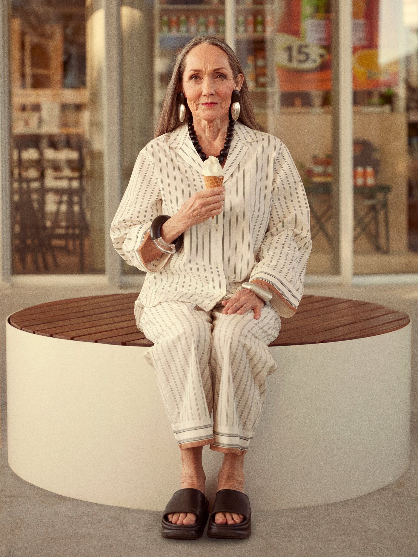 A woman wearing a white pyjama is sitting outside an IKEA store with a soft ice cream cone in her hand.