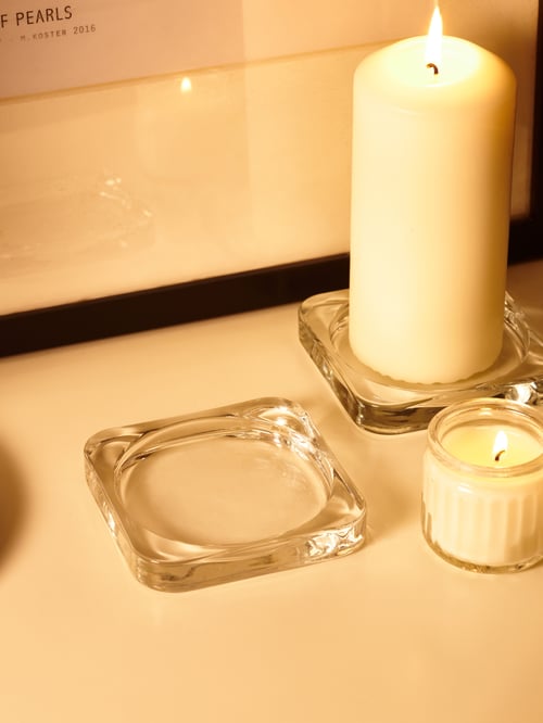 Eine FENOMEN Blockkerze steht hier neben einer ADLAD Duftkerze im Glas auf einer GLASIG Kerzen-/Dekoschale. 