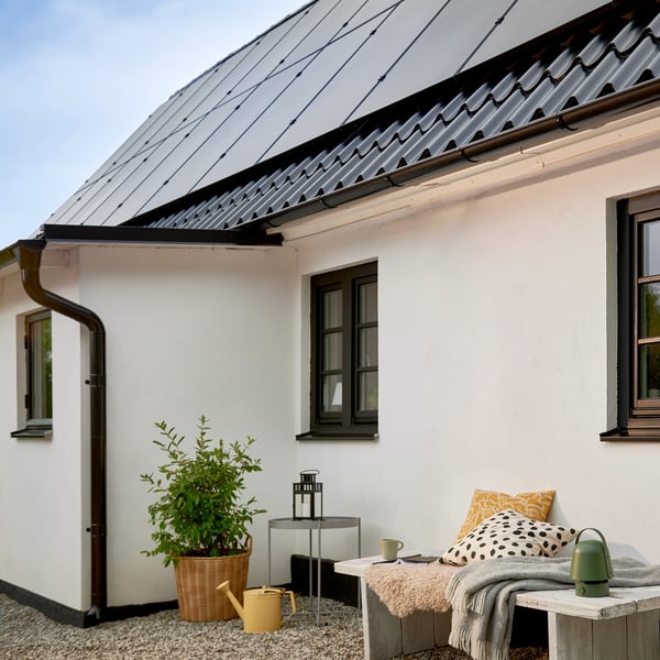 A charming white house with a black tiled roof featuring solar panels. We're looking at the side of the house where there is a small seating area.