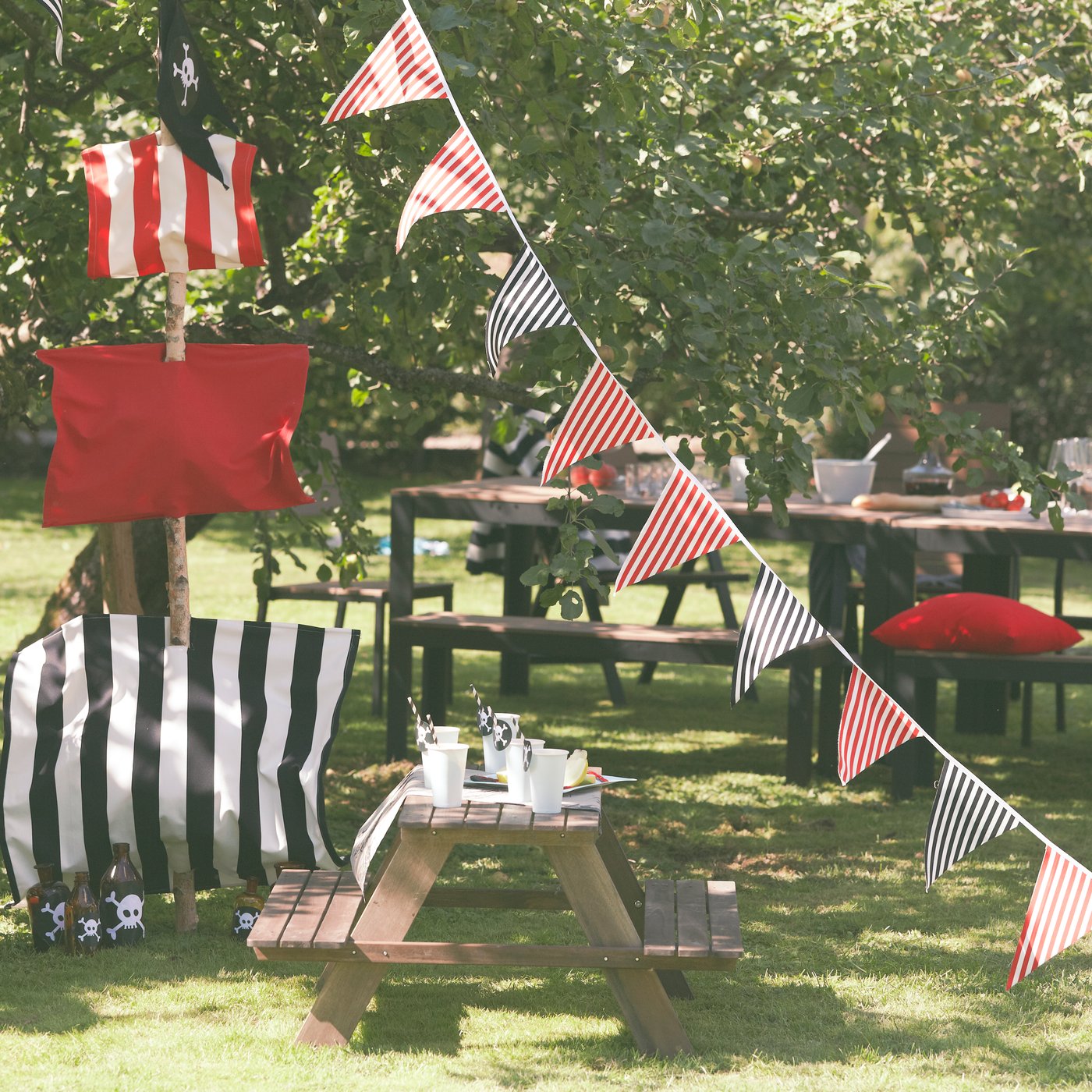 Outdoor picnic setup with striped bunting, picnic tables, and cushions under shady trees.