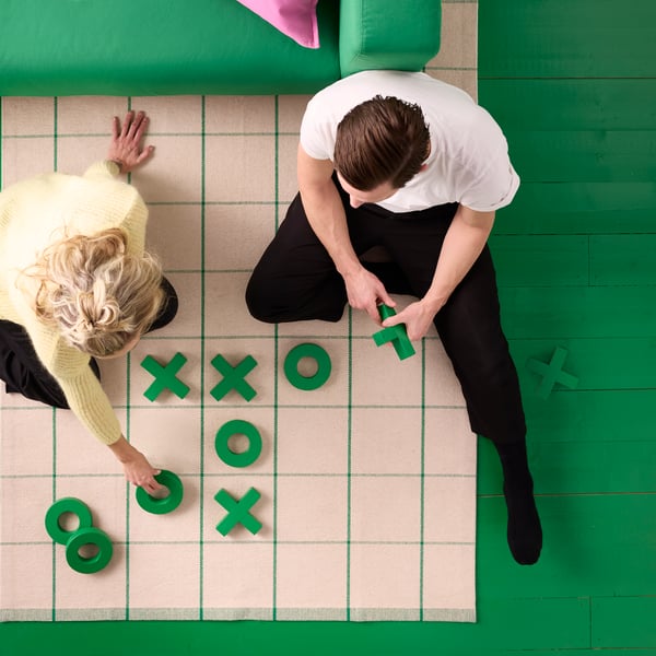 Two people are sitting by a green sofa on an off-white and green RIDSTIG flatwoven rug, playing noughts and crosses on it.