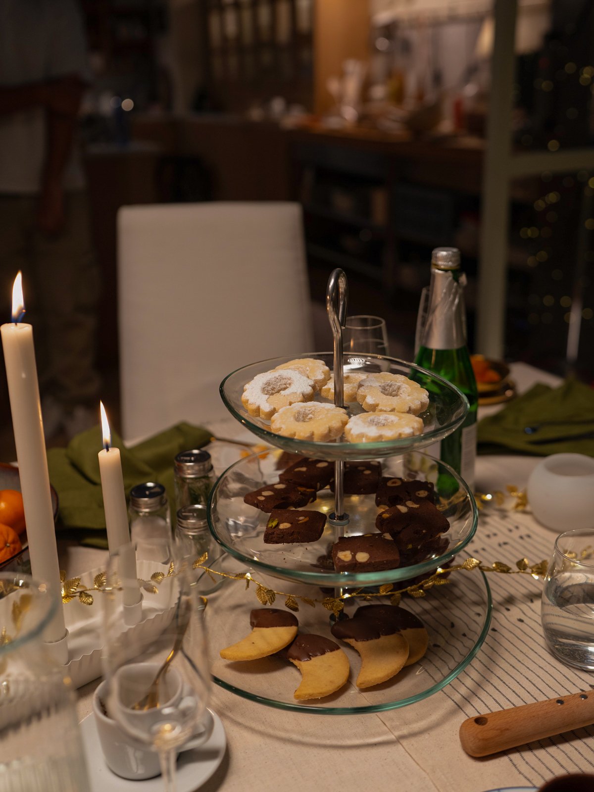 Un vassoio a due piani con biscotti decorati, posato su una tavola apparecchiata con candele bianche accese e bicchieri di vetro.
