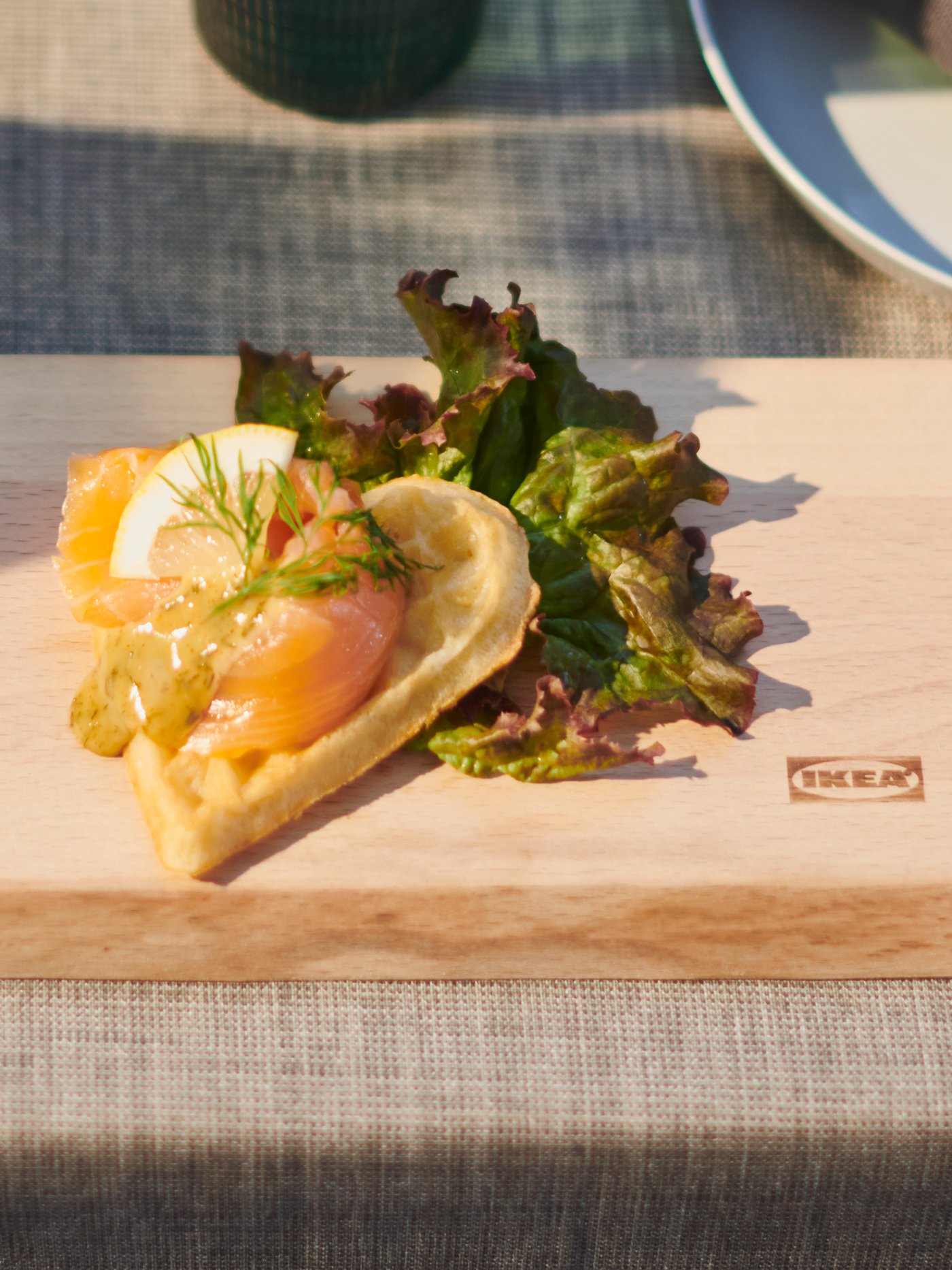 Una fetta di waffle su un tagliere di legno NÄBBFISK. Il waffle a la forma di cuore e una crosta dorata e croccante. Il condimento è a base di salmone affumicato e insalata. 