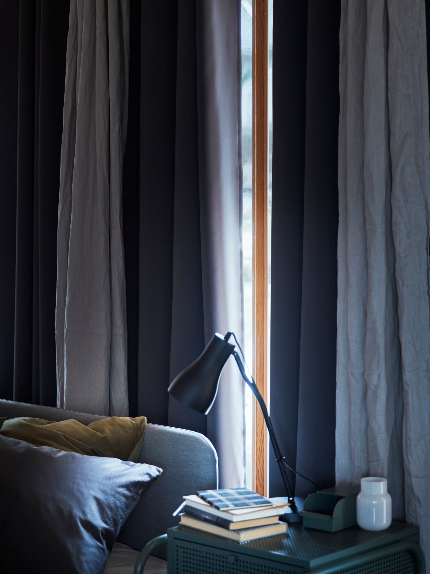 A night stand with a work lamp standing in front of a window with light linen curtains and dark HILLEBORG block-out curtains.