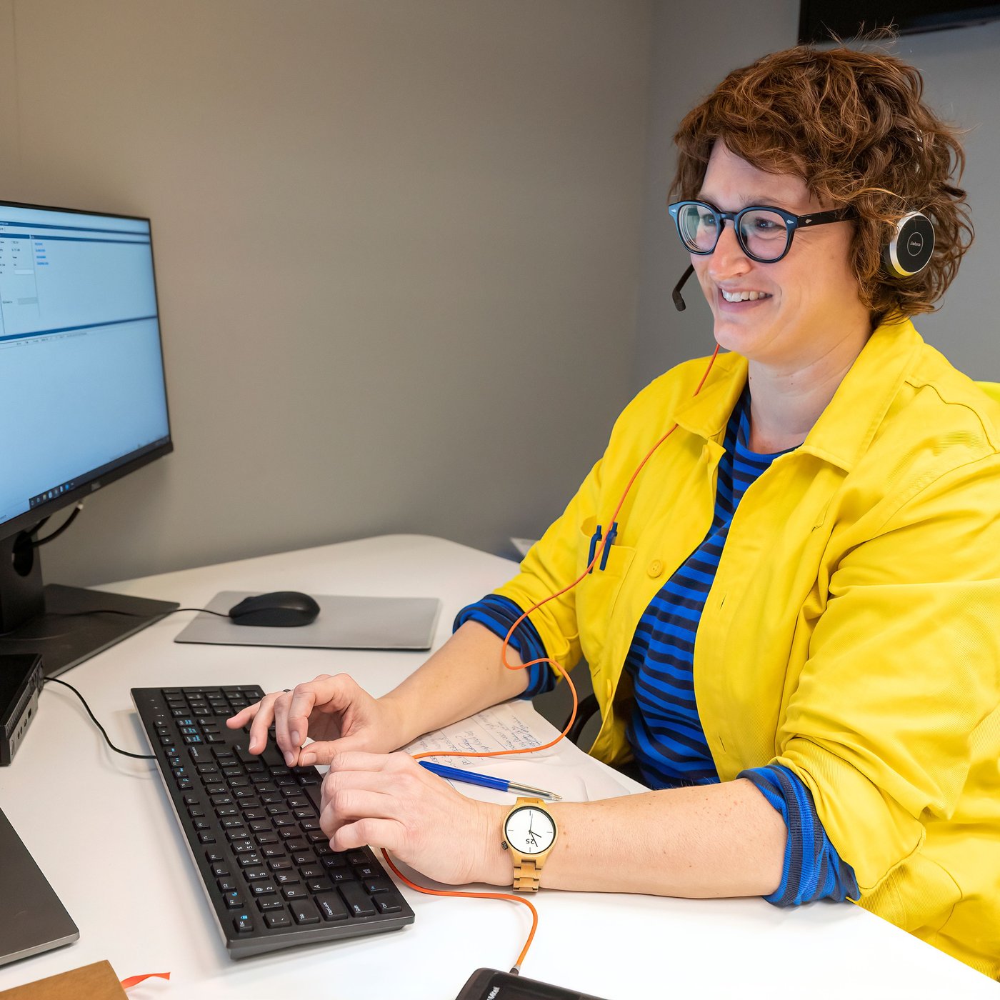 A smiling woman in blue-and-yellow IKEA co-worker clothing wearing glasses and a headset, sits at a desk looking at a screen.