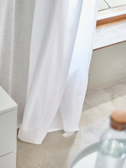 Bedroom close-up of a pair of white sheer curtains made of cotton and a defocused carafe with brown stopper.