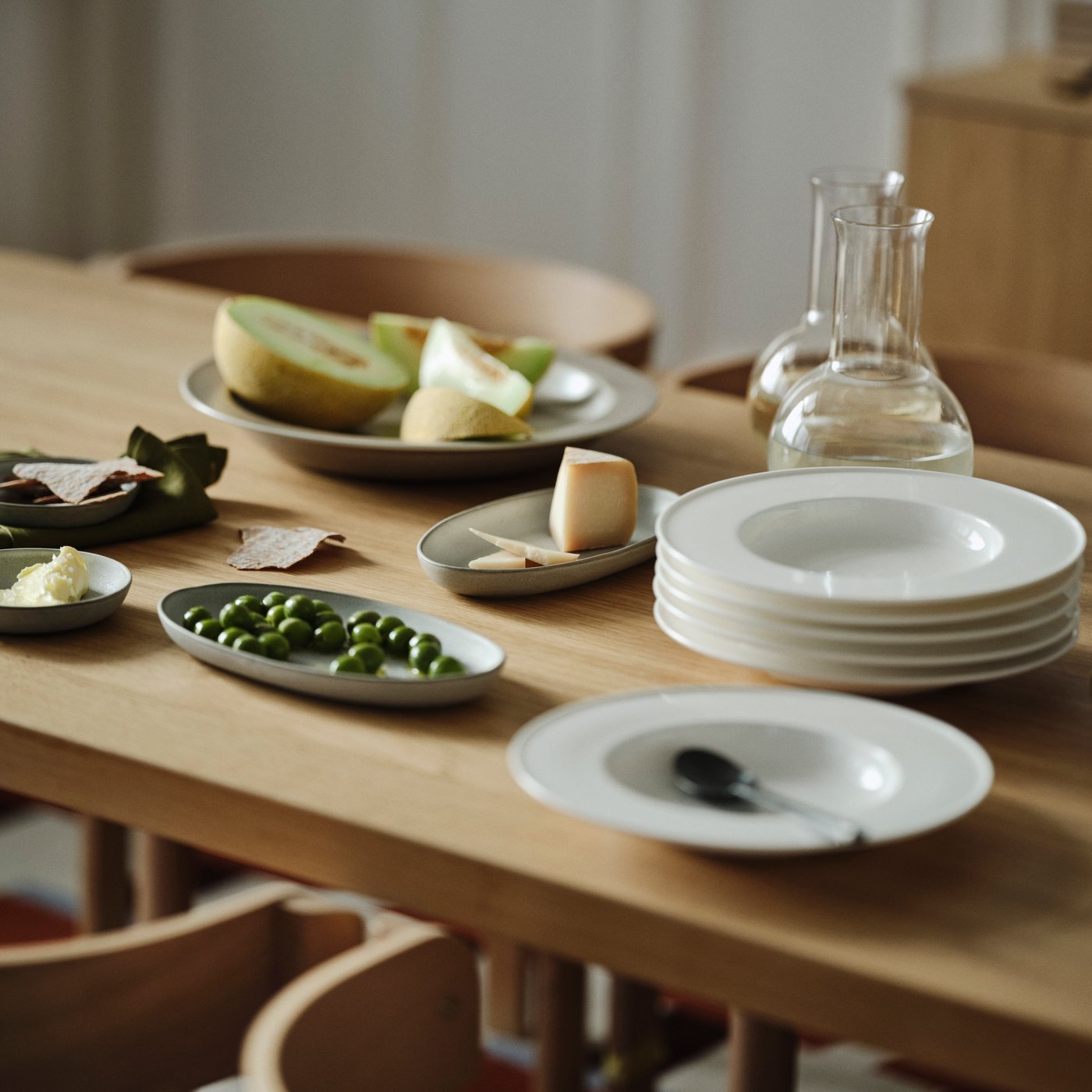 An oak veneer table with STOCKHOLM 2025 serving plates and serving bowls with food, carafes, and a stack of deep plates.
