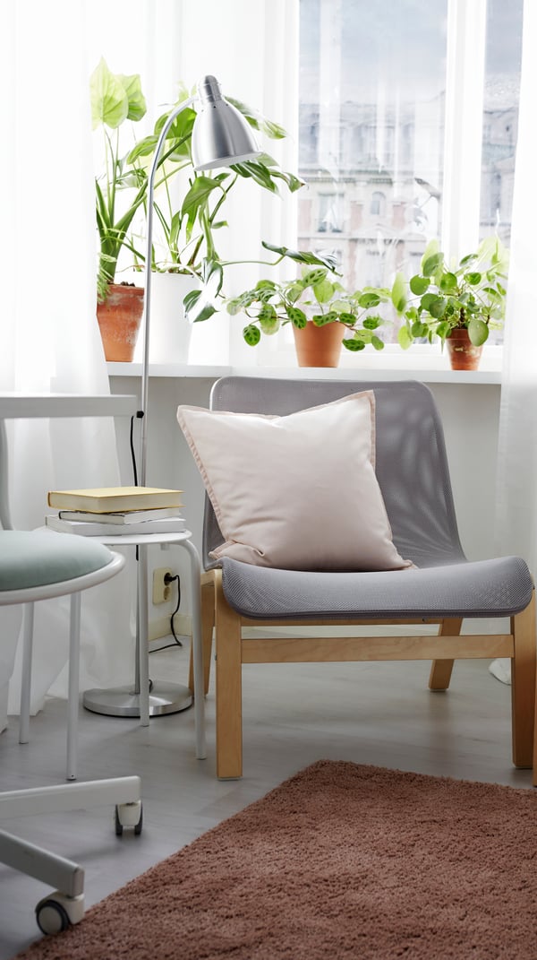 Poltrona NOLMYRA con impiallacciatura di betulla/grigio beige vicino ad una finestra con piante veri e su un tappeto marrone. Accanto c'è un tavolino coordinato.