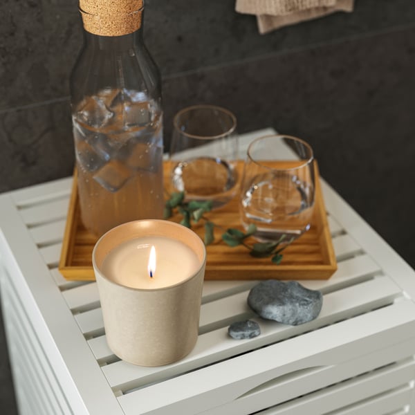 The top of a MUSKAN storage stool with a tray holding a bottle and glasses of water and a lit JÄMLIK scented candle on it.