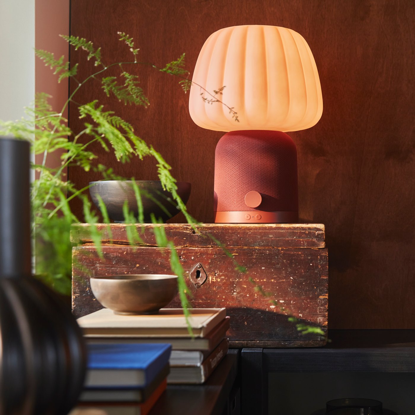 A KULGLASS Bluetooth speaker lamp in red brown/pink is placed on a wooden box surrounded by books, bowls and a plant.