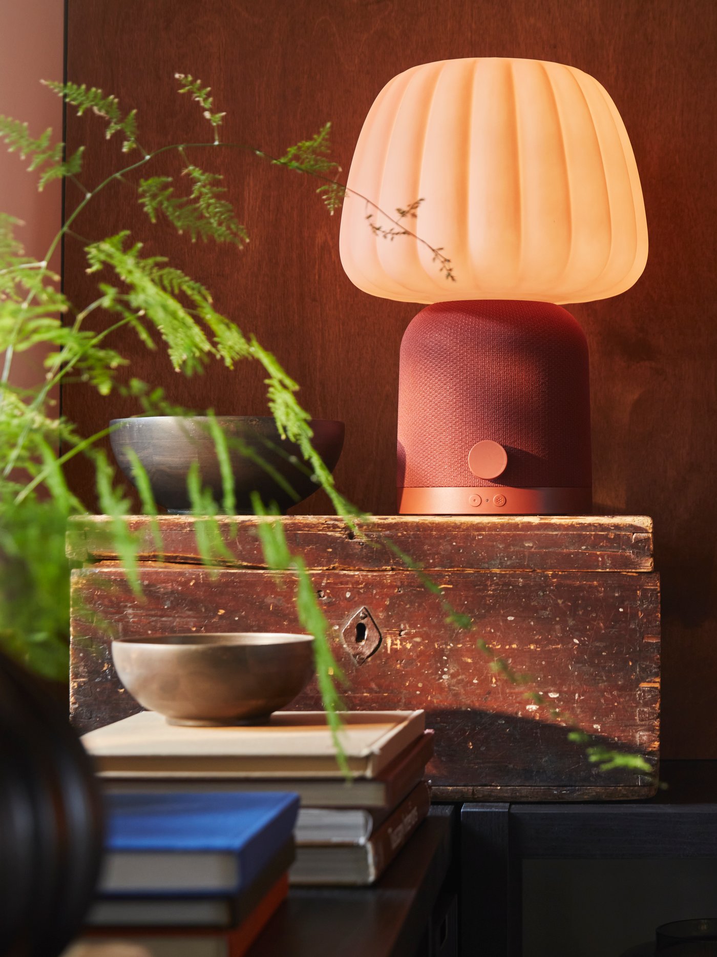 A KULGLASS Bluetooth speaker lamp in red brown/pink is placed on a wooden box surrounded by books, bowls and a plant.