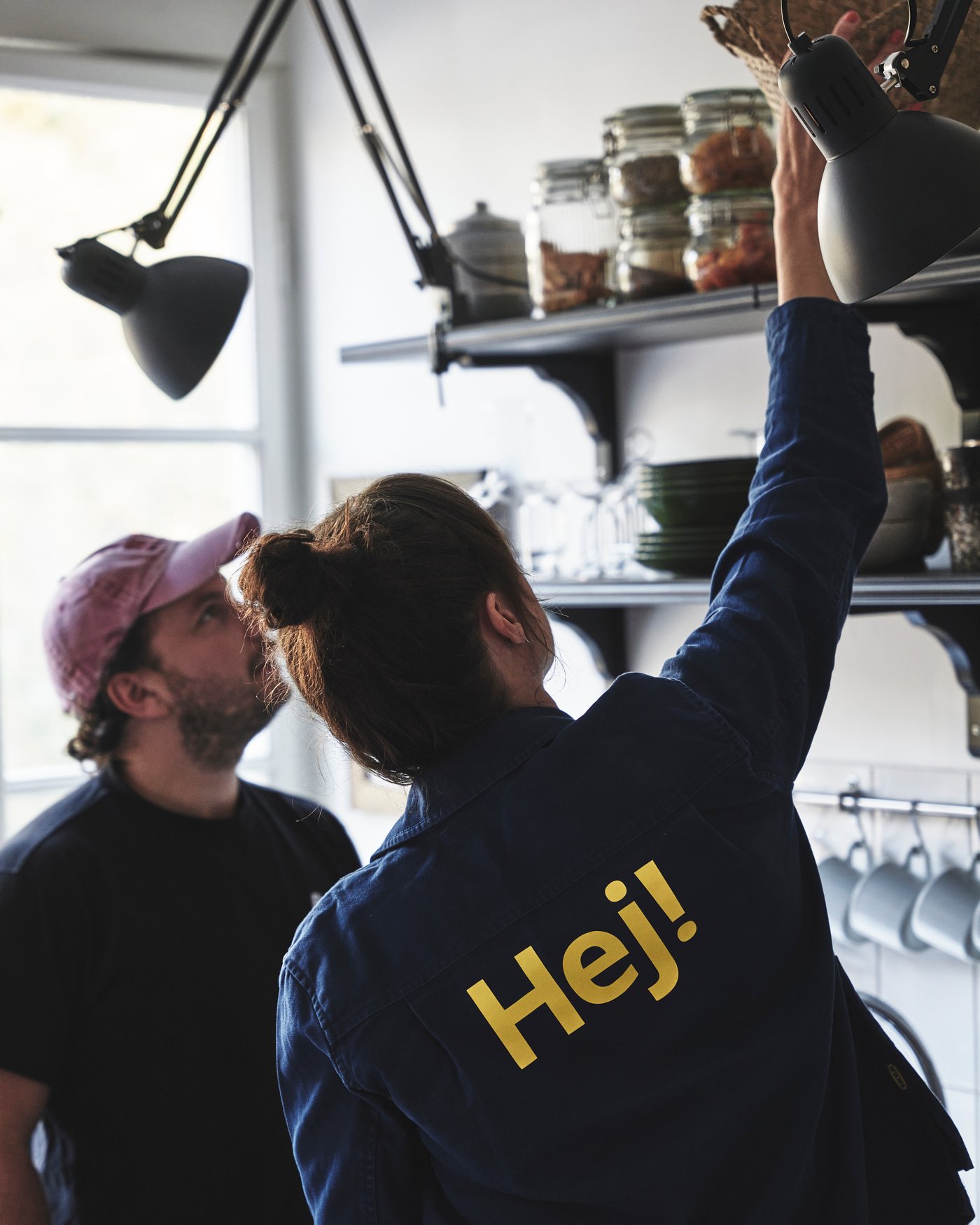 En IKEA medarbetare sträcker sig för att lägga en korg på en svart hylla. I bakgrunden syns en man med röd keps.