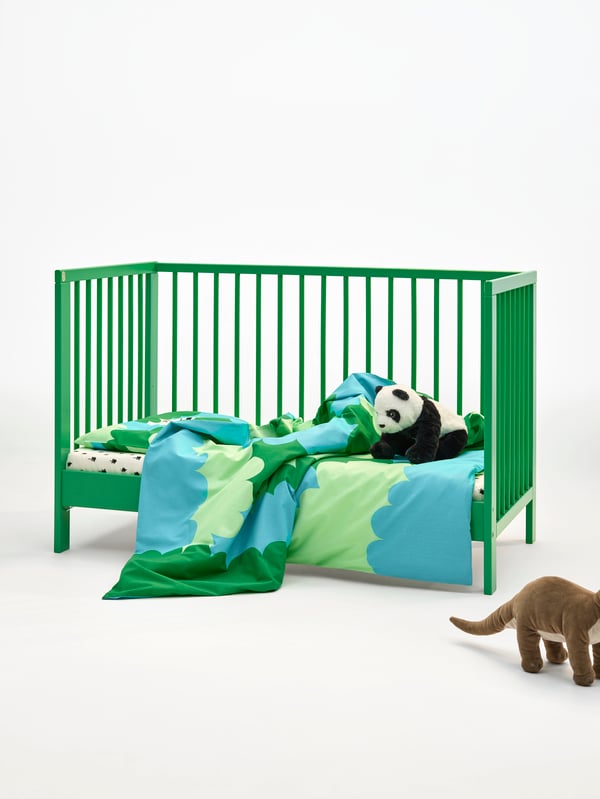 A green cot with colourful bedding in a white space. The side of the cot is open so it can be used as a child’s bed.