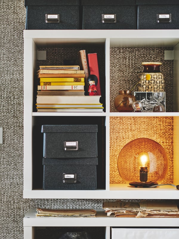 Close up on a KALLAX bookshelf placed against a wall. There are several TJOG boxes, books and a FADO lamp on the shelves.