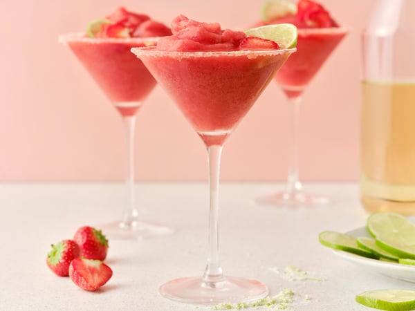 Three tall glasses filled with ISKUB elderflower soft drink and frozen strawberry margaritas, garnished with lime.