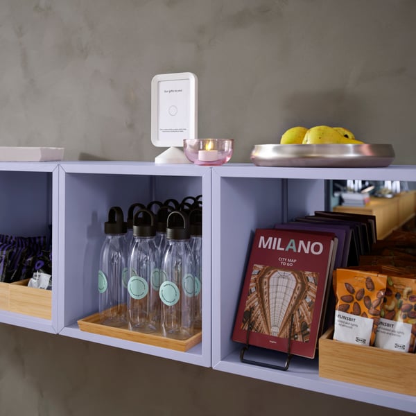 A row of three pale lilac EKET cabinets are mounted on a cement wall. They hold tourist maps, water bottles and snacks.