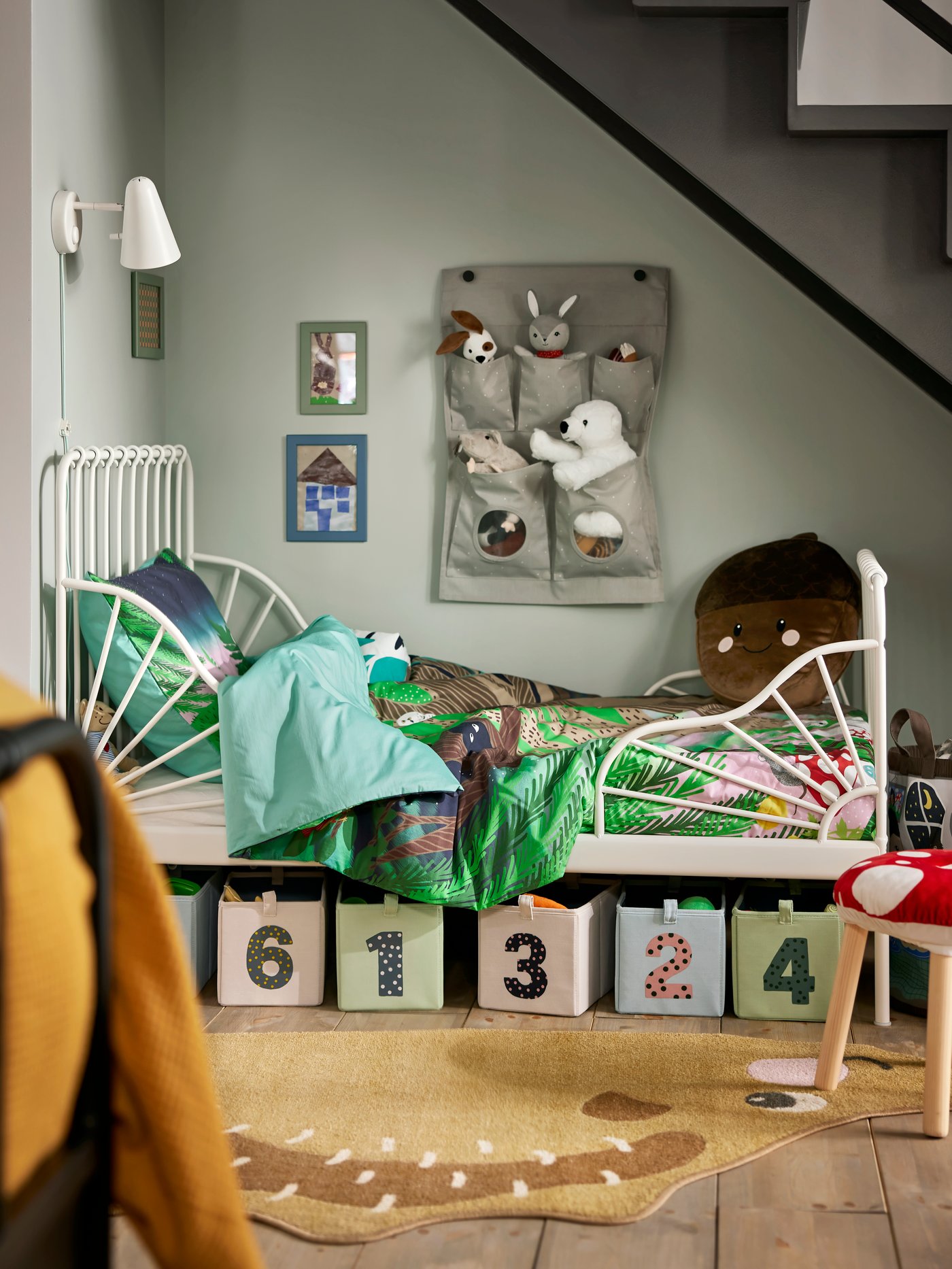Kinderbett mit grün gemusterter Bettwäsche und nummerierten Aufbewahrungsboxen darunter. An der Wand hängen dekorative Elemente und ein Plüschtier. Helle, freundliche Atmosphäre mit Holzfußboden.