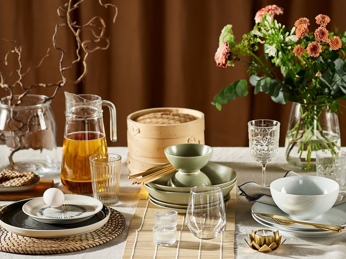 Es ist ein Esstisch mit vielen Gegenständen zu sehen, u. a. Blumen, Gläser, FÄRGKLAR Teller in mattiertem Grün und eine TILLBRINGARE Kanne.