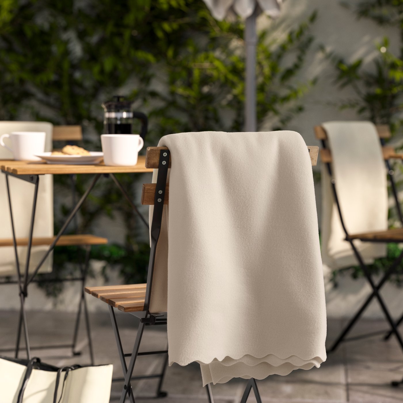 Ein DAGGAKÅPOR Plaid in Cremeweiß liegt dekorativ über der Rückenlehne eines Outdoor-Stuhls. Die weiche Decke mit gewelltem Rand verleiht dem Essplatz im Freien eine gemütliche, einladende Atmosphäre.