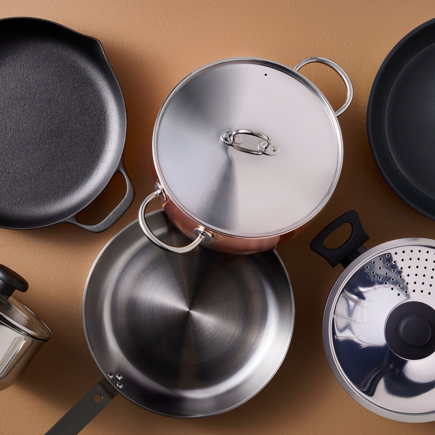 Pots and pans made of different materials, from cast iron to stainless steel, are placed on a light brown surface.