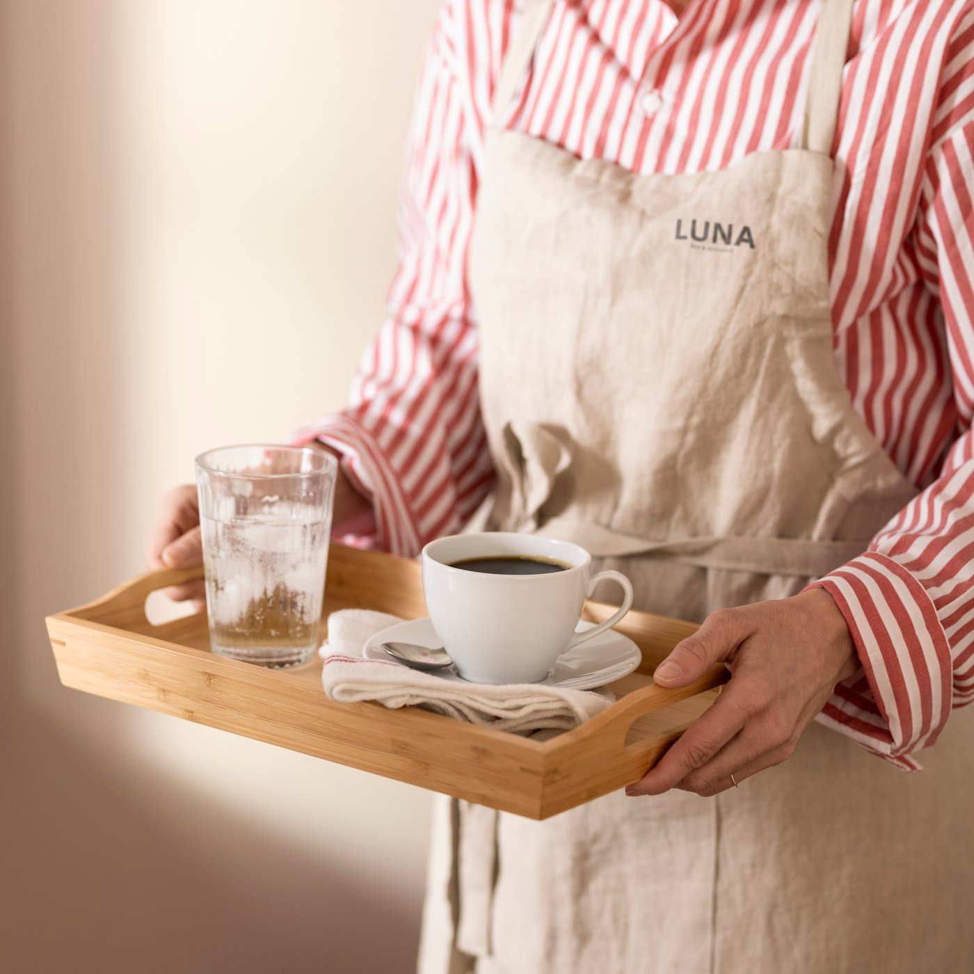 Una persona che indossa un grembiule tiene in mano un vassoio rettangolare di bambù FRUKTSKAL con sopra un bicchiere di acqua ghiacciata e una tazza di caffè.