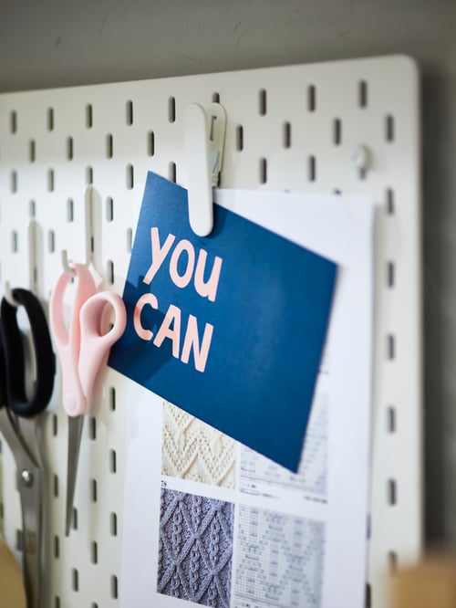 A white SKÅDIS pegboard with various notes and scissors hanging on hooks and a clip holding paper.