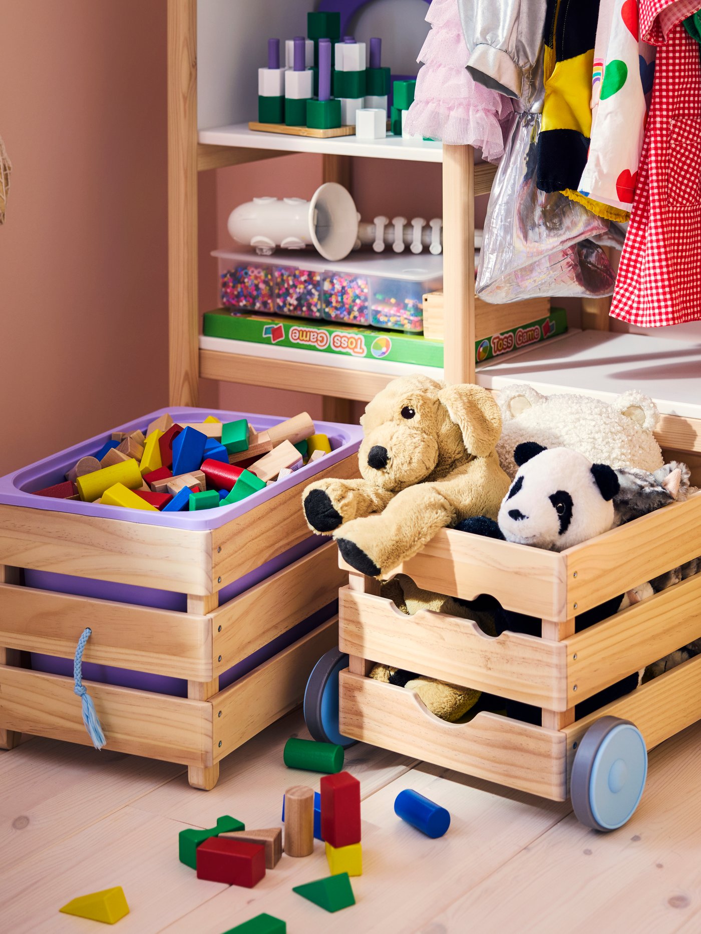 Two FLISAT toy storage with wheels in pine, filled with building blocks and soft toys, in a pink children’s room.