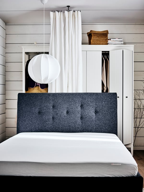 A white bedroom with an IDANÄS upholstered bed with no bed linen on, in front of two white IDANÄS wardrobes and a curtain.