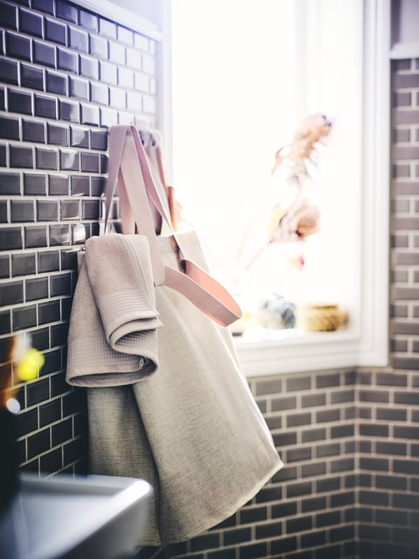 A bathroom with black tiled walls, featuring a PURRPINGLA laundry bag hanging on hooks, and containing a VINARN bath towel.