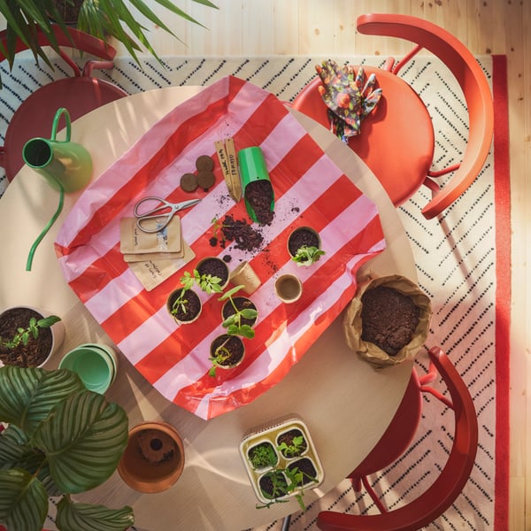 A bright pink and red striped DOFTRIPS potting mat placed on a round table filled with small pots, seedlings and tools.
