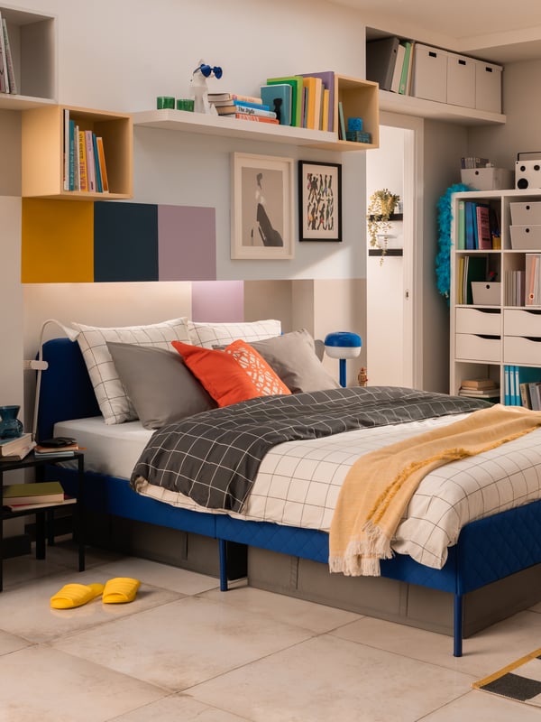 A well-organized bedroom featuring a blue bed with gray and white bedding, accented by an orange pillow. A beige throw is draped over the foot of the bed. The room has white walls with a series of colorful square and rectangular panels above the bed, alongside framed artwork. A shelving unit to the left of the bed holds books, storage boxes, and decorative items. To the right, there is a white cabinet with more books on top. Yellow slippers are placed neatly on the floor beside the bed.