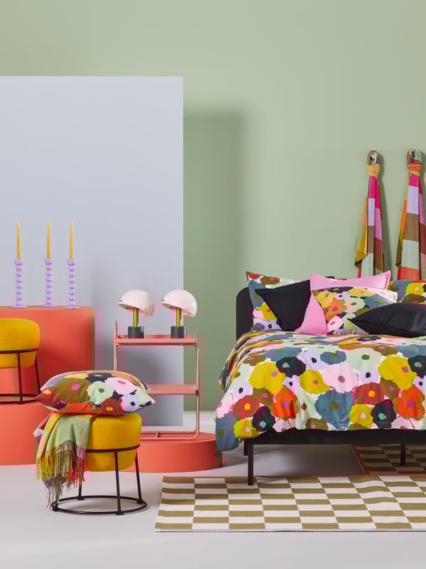 Una cama vestida con una funda nórdica MURREVA, rodeada por un puf EMMISLÖV amarillo y una mesilla de noche/estantería HATTÅSEN rosa.