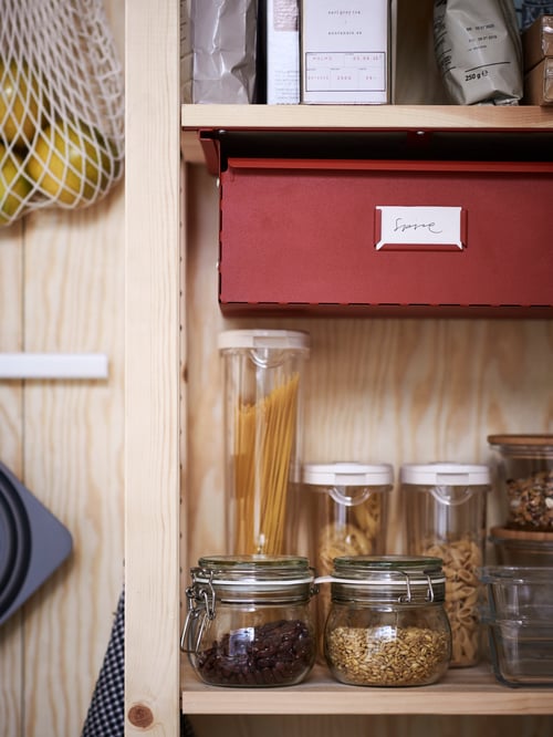 Open, pine shelves hold clear glass KORKEN jars with lids containing dried beans and grains, below a red metal spice drawer.