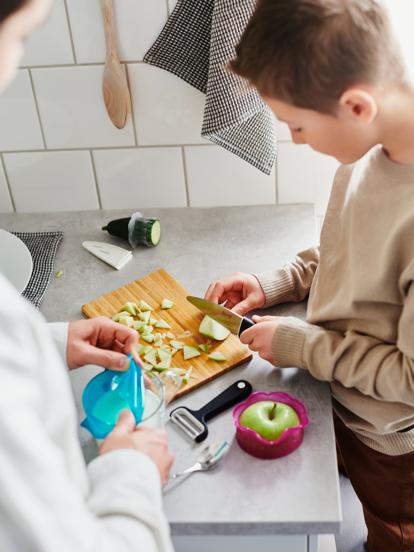 A boy stands by a worktop, focusing on dicing an apple with a SMÅBIT knife on a chopping board, with a SMÅBIT peeler beside.
