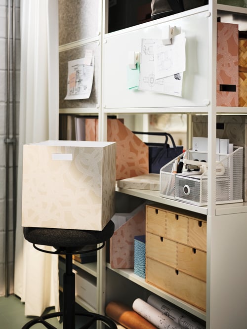Une boîte de rangement TJENA à motif beige est posée sur un tabouret à côté d’une étagère blanche.
