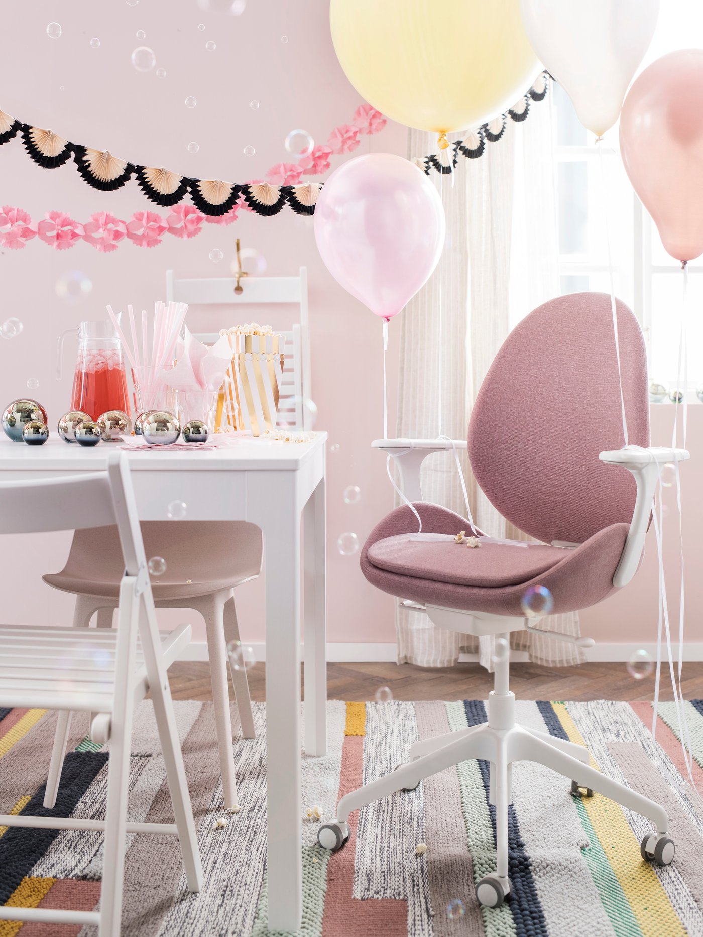 En lyserrød kontorstol står for enden af et bord og der hænger forkelligfarvede helliumballoner fast i. Der er pyntet op til fest med gurilander og drinks på bordet. 