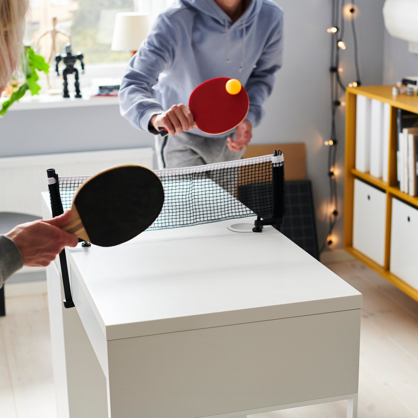 Niños jugando ping pong en una mesa blanca MICKE. Al costado de la habitación hay luces led colgantes VISSVASS y una estantería de color mostaza EKET.