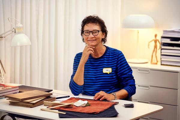 En interiørdesigner fra IKEA i en blå genser sitter ved et skrivebord med interiørprøver og smiler til kameraet.