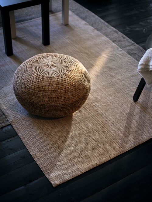 O pouffe bej așezată pe un covor dreptunghiular, cu țesătură plată. Picioarele scaunului sunt vizibile la ambele capete ale covorului.