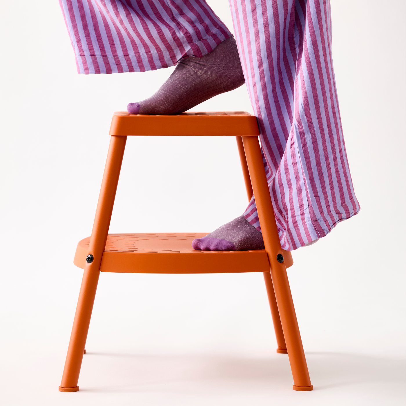 A person in purple and white striped trousers and purple socks is stepping onto a HADDARP step stool in 4-way orange.