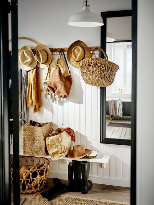 A white wood-panelled hallway with a row of hooks holding straw hats and baskets. A bench is below and a mirror beside it.