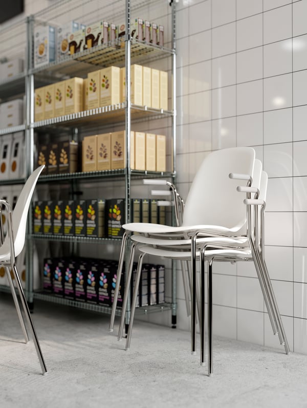 Three LEIFARNE white chairs stacked together, shown next to an open shelving unit filled with food packages.
