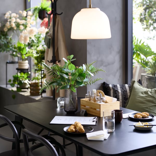 Close-up of a café  with a black LISABO table, served food, a grey vase with greenery and flowers in the background.