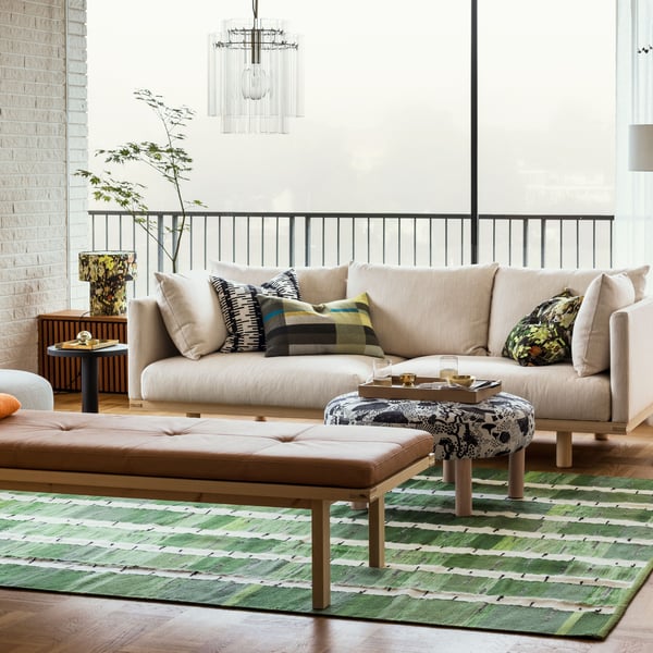 Un luminoso soggiorno con un divano beige a tre posti STOCKHOLM 2025, una panca e un pouf appoggiati su un tappeto verde, nero e bianco.