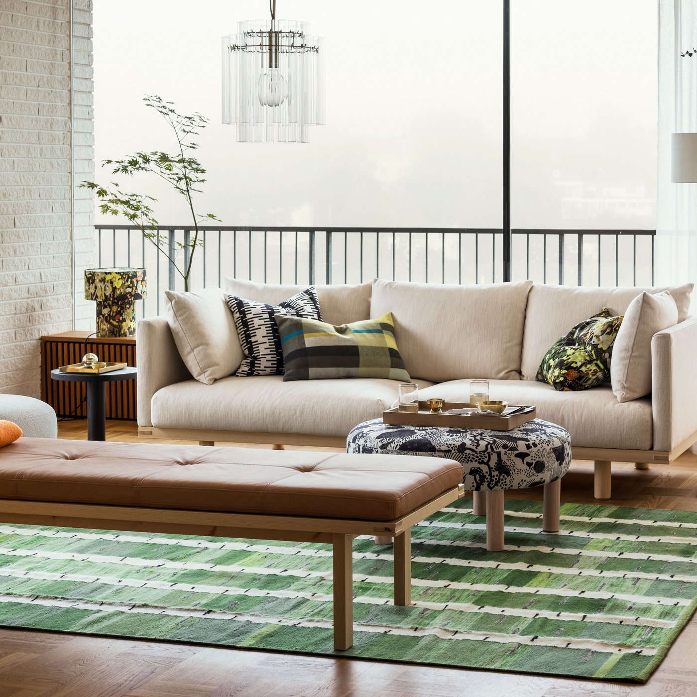 Un luminoso soggiorno con un divano beige a tre posti STOCKHOLM 2025, una panca e un pouf appoggiati su un tappeto verde, nero e bianco.